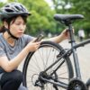 公園で、ヘルメットをかぶった女性が携帯工具を手に、動かなくなった自転車のサドルレバーを困った表情で見つめている様子。
