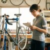 整備された工房で、クラシックなリムブレーキのロードバイクの横に立ち、メンテナンス計画のノートを見ながら思案する若い日本人女性。愛車を10年乗り続けるための戦略と維持管理を象徴するシーン。